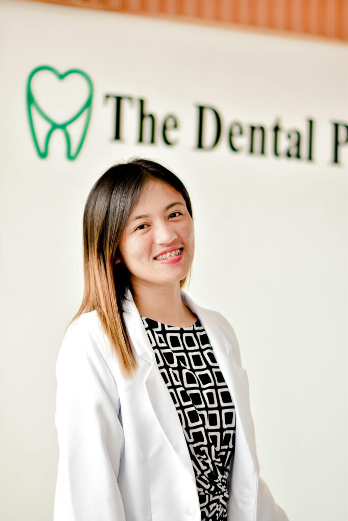 A confident female dentist in a white coat smiling warmly against a dental clinic backdrop.
